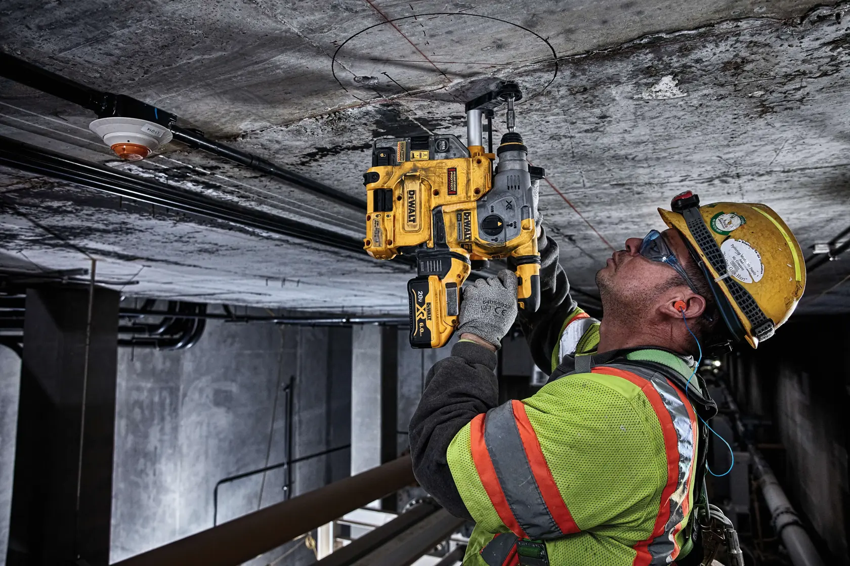 A construction worker wearing a safety helmet and reflective vest is using a DEWALT rotary hammer drill to drill into a concrete ceiling in an industrial setting.