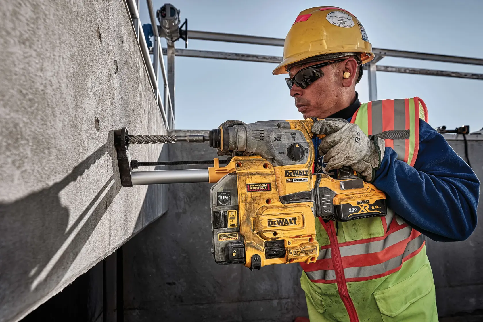 A construction worker wearing a safety vest, gloves, and a hard hat operates a DEWALT DCH293R2 rotary hammer drill, drilling into a concrete wall at a worksite.