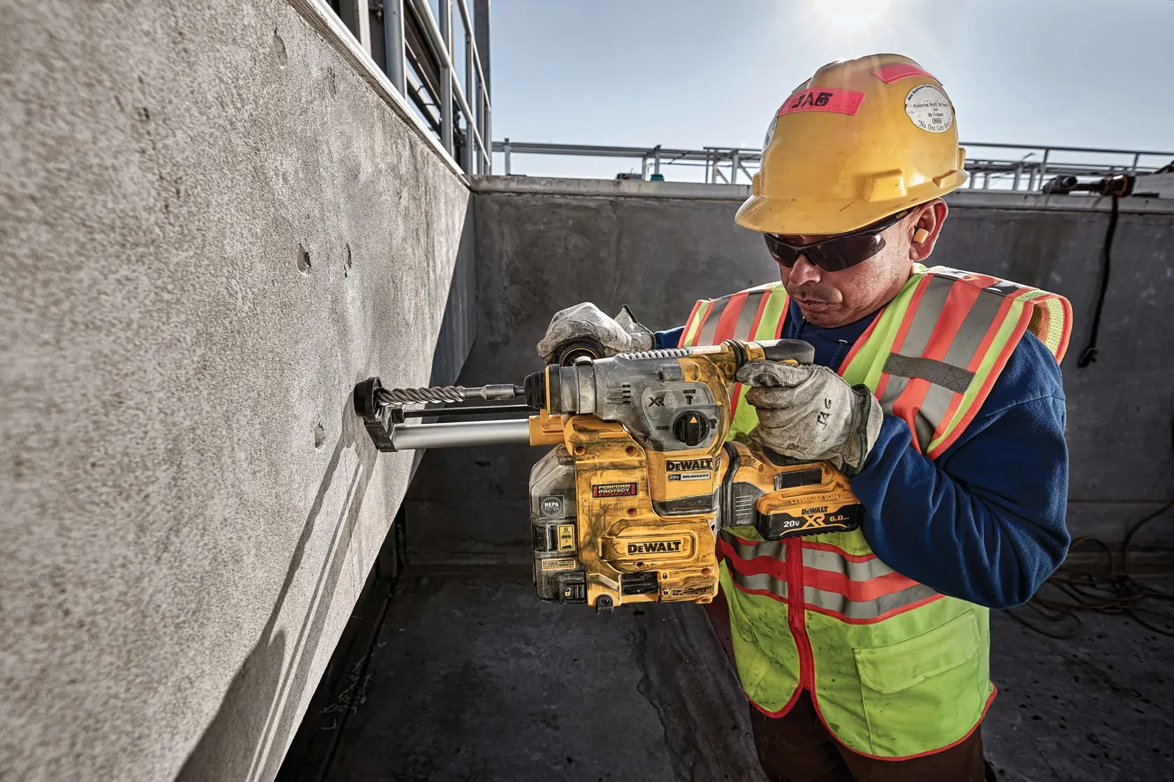 A person wearing a yellow hard hat, safety gloves, and a reflective vest operates a DEWALT cordless rotary hammer drill against a concrete wall at a construction site.