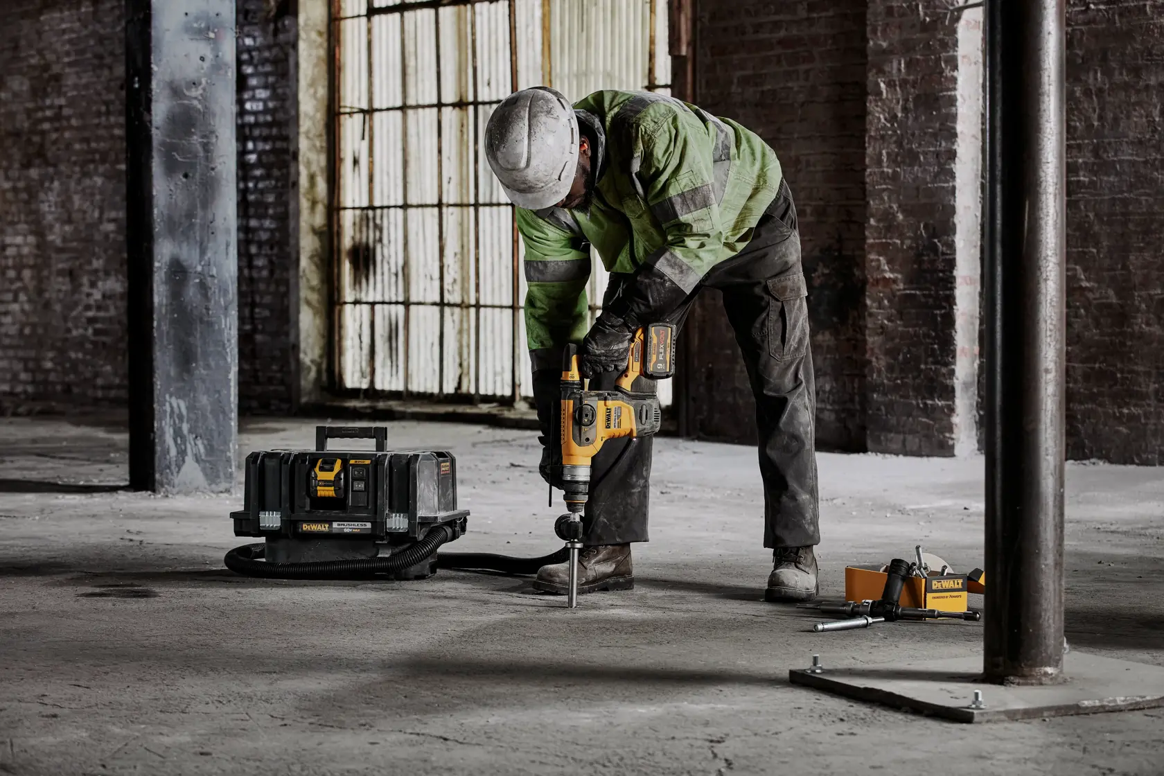 A worker wearing a helmet and safety gear is using a DEWALT tool to drill into a concrete floor in an industrial setting. A DEWALT toolbox and other equipment are placed nearby.
