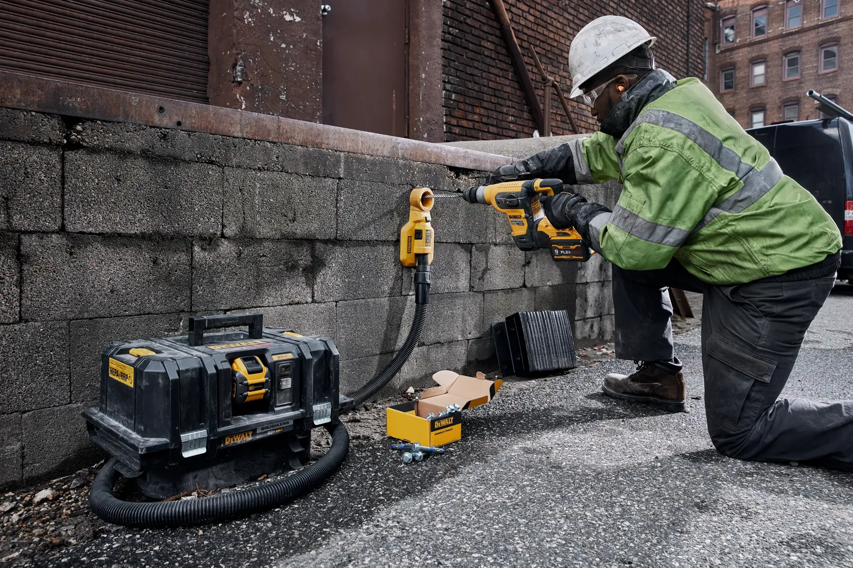 A construction worker wearing safety gear is kneeling on the ground and using a DEWALT rotary hammer to drill into a concrete wall. The worker is connected to a DEWALT dust extraction system and there is a DEWALT storage case and a box of accessories nearby.