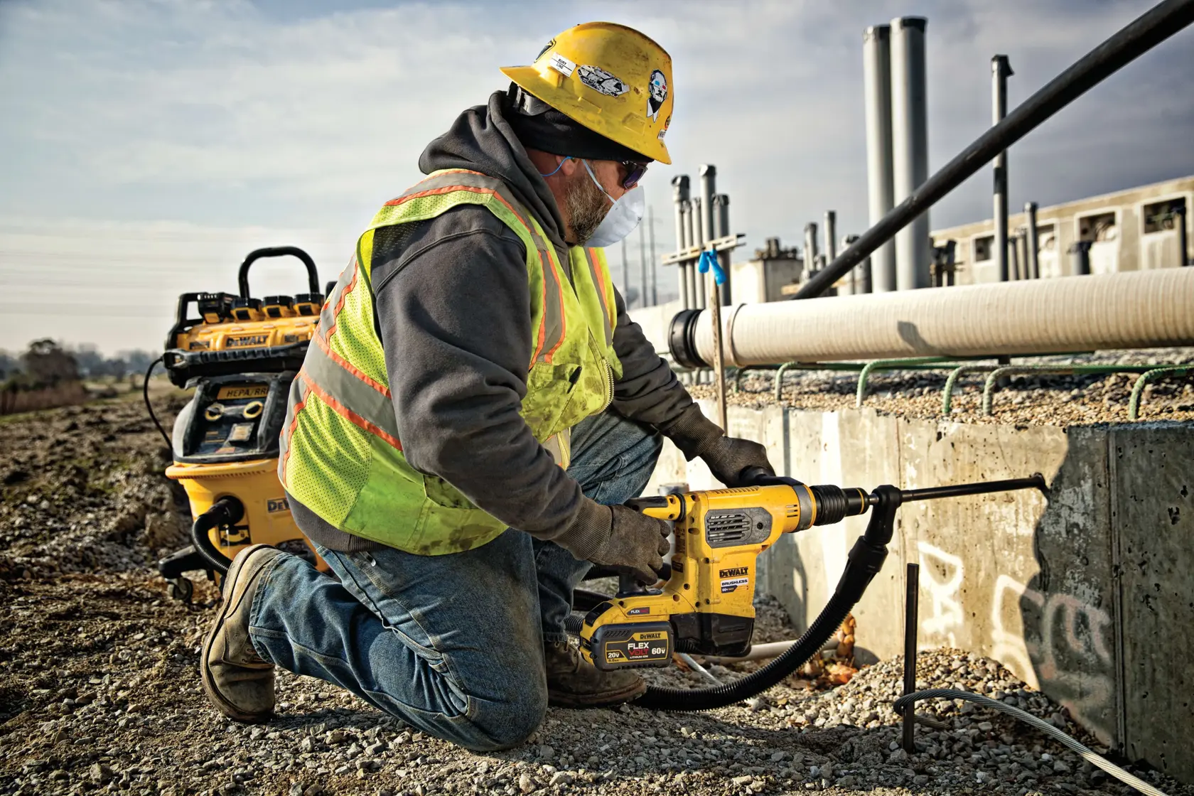 A construction worker wearing a yellow hard hat and safety vest is kneeling on gravel while using a DEWALT power tool to drill into a concrete wall at an outdoor construction site. The worker's face is blurred for privacy.