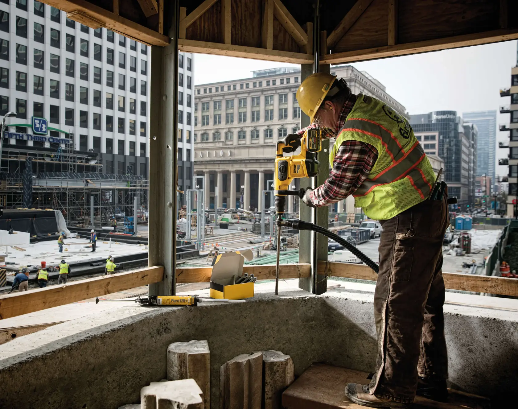 A construction worker wearing a safety vest and helmet is using a DEWALT power tool to drill into a concrete structure at a building site in an urban area. Several other workers are visible in the background amongst scaffolding and buildings.