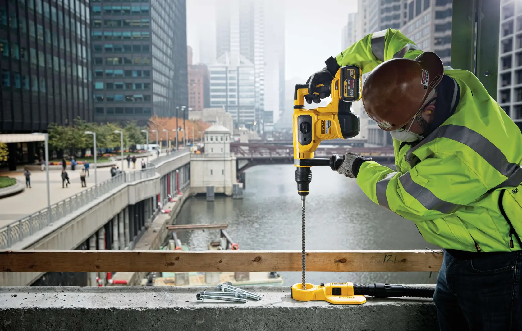 A construction worker wearing a safety helmet and high-visibility jacket uses a DEWALT rotary hammer drill (SKU: DCH481X2) to drill into concrete on a city building site next to a river with skyscrapers in the background.