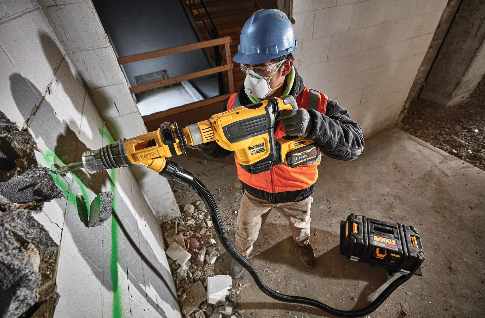 A construction worker wearing a blue hard hat and orange safety vest uses a DEWALT rotary hammer drill (model DCH614B) to break through a concrete wall. The tool is connected to a dust extraction system, and a DEWALT tool case is placed on the ground nearby.
