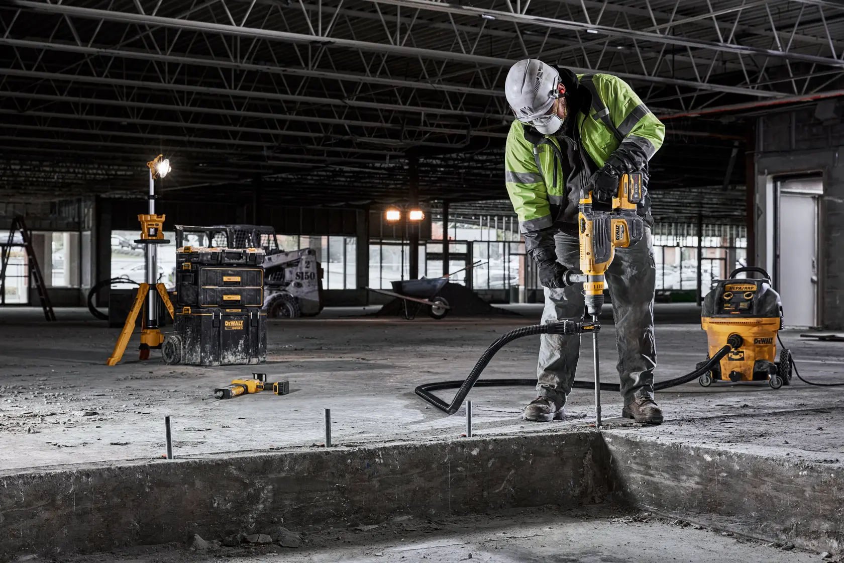 A construction worker wearing safety gear uses a DEWALT power tool to drill into concrete inside an unfinished building. Various DEWALT tools and equipment are visible around the work area.