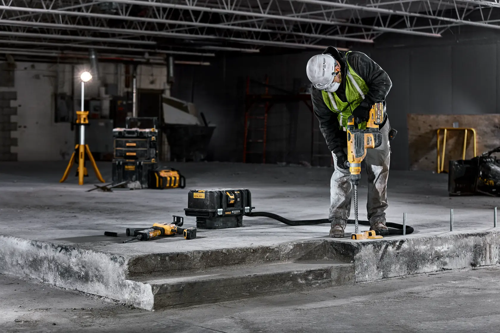 A worker wearing a safety vest and helmet uses a DEWALT cordless rotary hammer drill to break concrete indoors. DEWALT power tools and equipment, including a light and tool cases, are visible in the background.