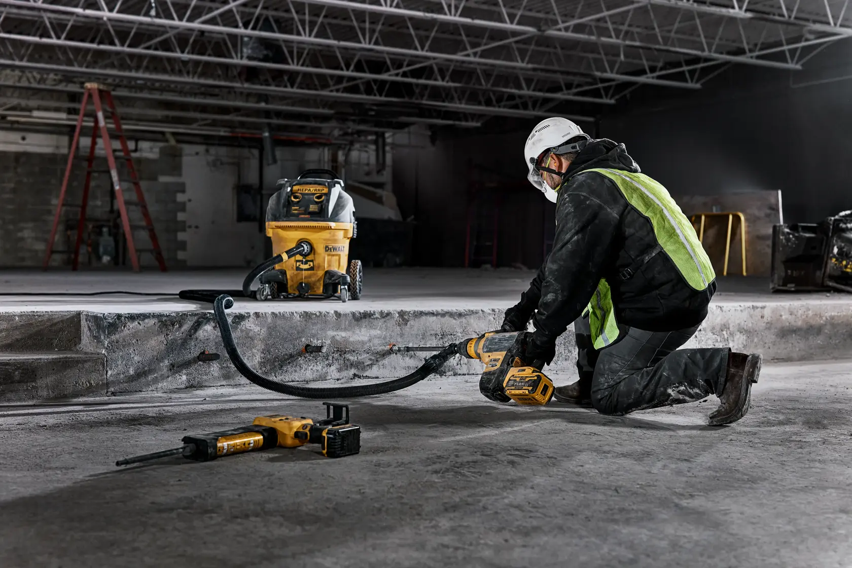 A construction worker wearing a white helmet, safety glasses, and a high-visibility vest is kneeling on a concrete floor indoors while using a yellow DEWALT power tool, attached by a hose to a yellow DEWALT vacuum. Another DEWALT power tool lies on the ground nearby in the industrial setting.