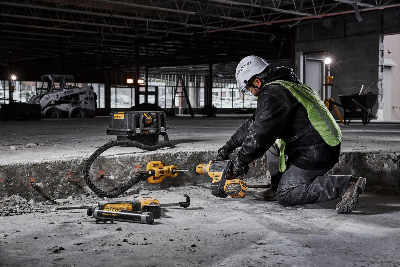 A construction worker wearing a safety helmet and high-visibility vest is kneeling on the ground and using a DEWALT cordless hammer drill to break concrete indoors. Several DEWALT power tools and accessories are placed nearby on the floor. The worker is in an unfinished building with construction equipment visible in the background.