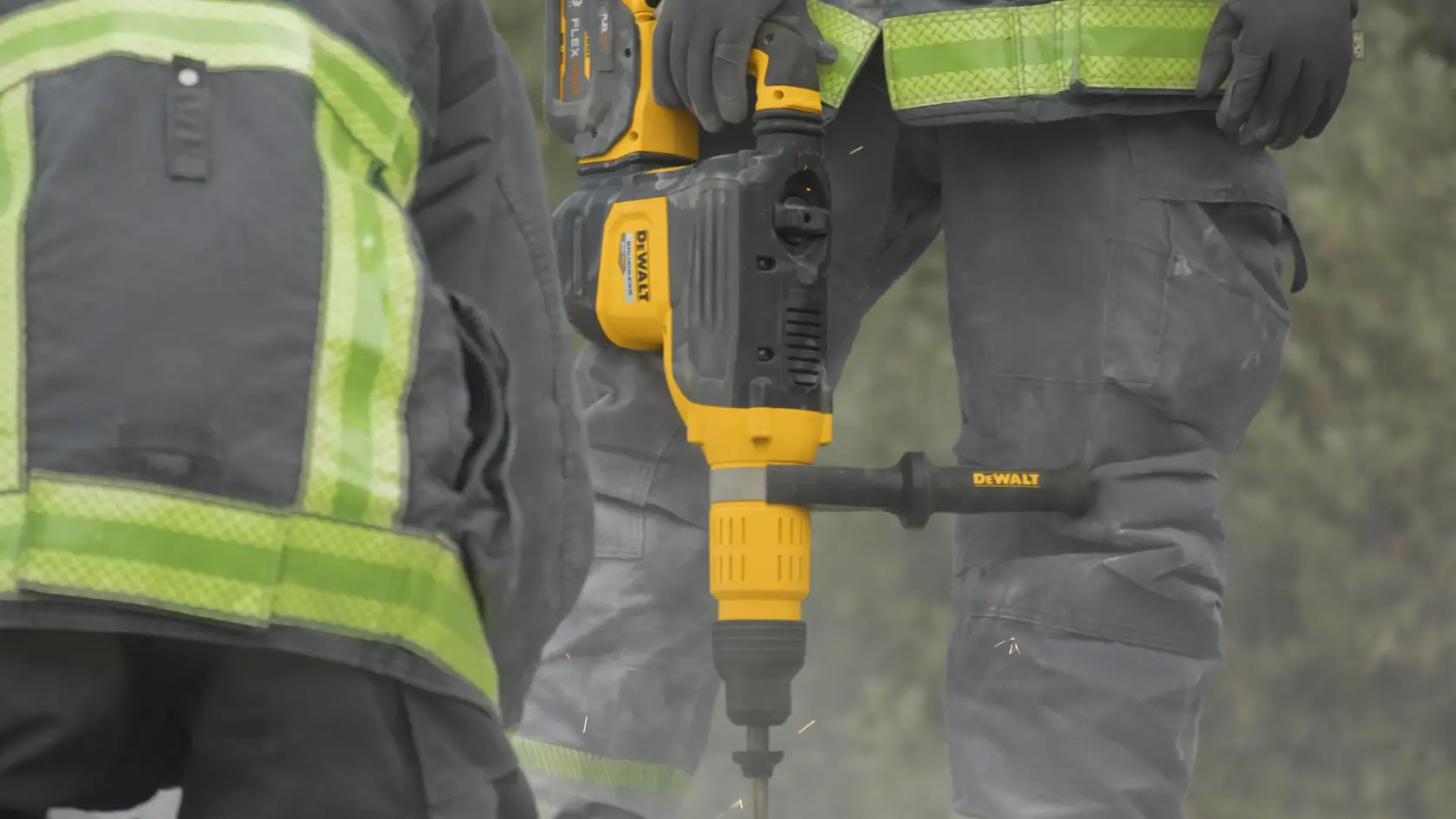 Two workers wearing safety vests and gloves are using a DEWALT DCH775X2 drill on a construction site. The yellow and black power tool is held by one worker and is actively drilling into the ground.