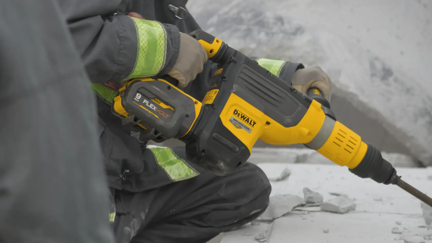 A person using a yellow and black DEWALT cordless rotary hammer drill to break concrete outdoors. The tool has FLEXVOLT battery technology.