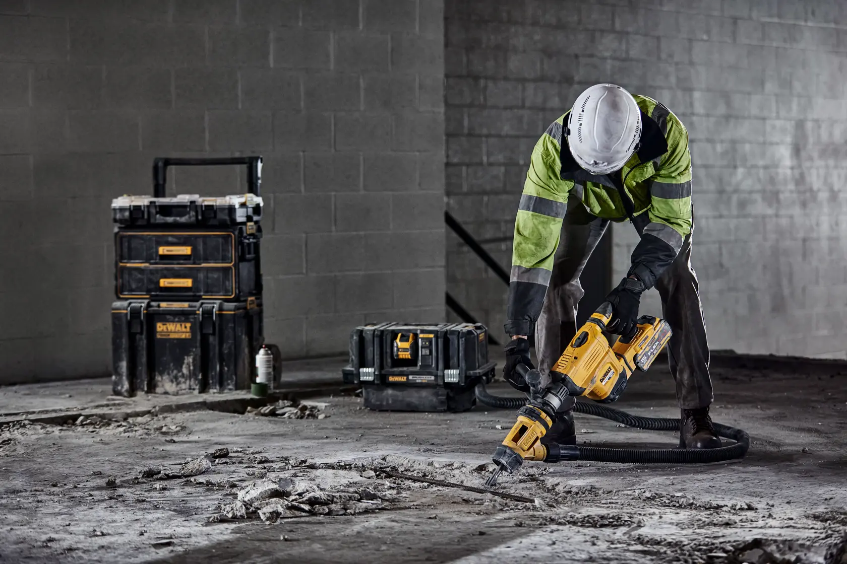 A construction worker wearing a safety jacket and helmet uses a yellow DEWALT demolition hammer to break up concrete on a building site. DEWALT toolboxes are visible in the background.
