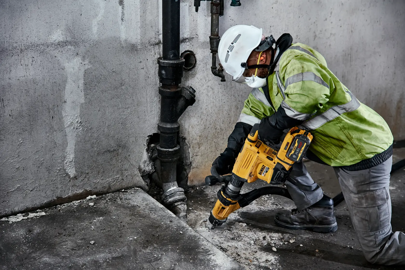 A construction worker wearing safety gear uses a yellow DEWALT demolition hammer to break concrete near a wall and pipe.