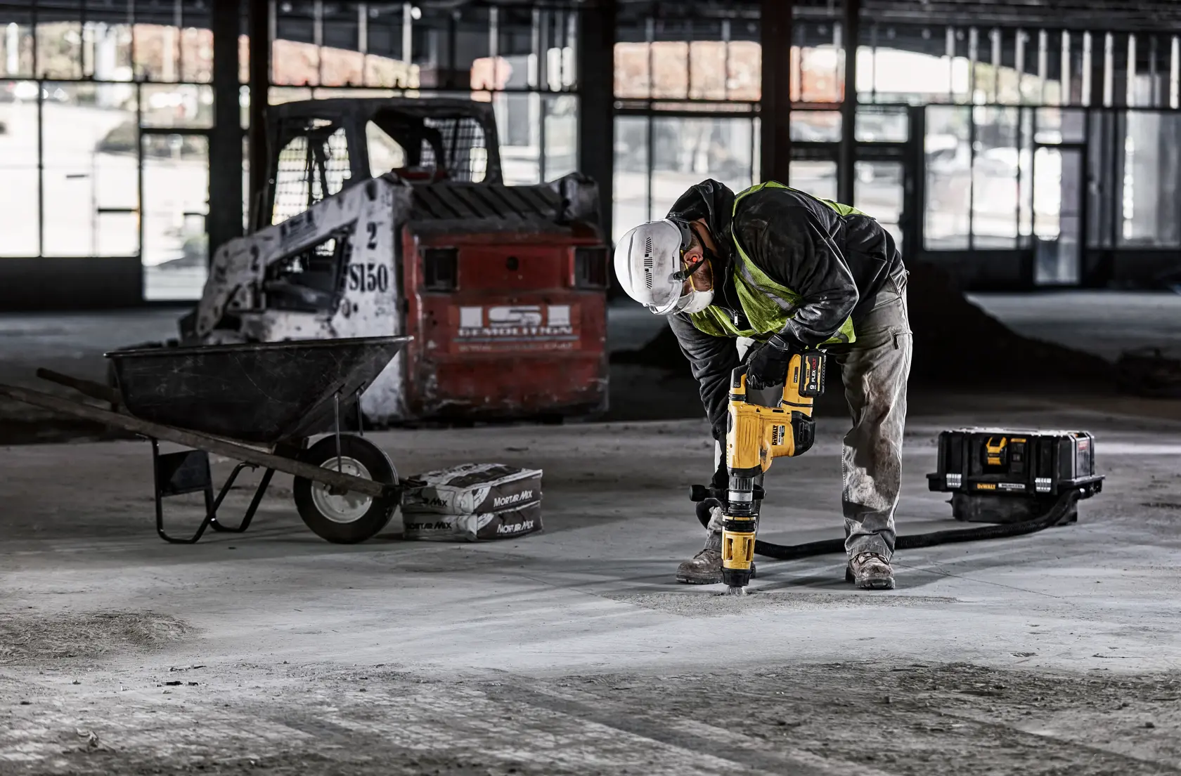 A construction worker wearing a hard hat and safety vest uses a yellow DEWALT power tool to drill into a concrete floor inside an unfinished building. A wheelbarrow, a toolbox, and construction equipment are visible in the background.