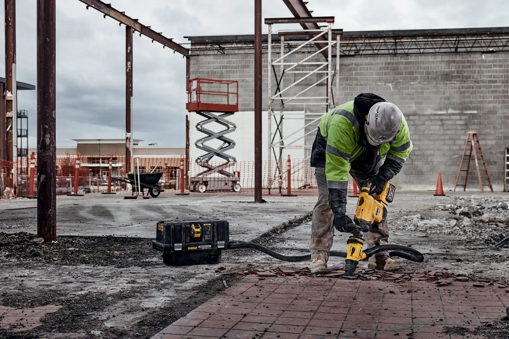 A construction worker wearing safety gear uses a yellow DEWALT rotary hammer drill to break up a brick surface at an outdoor construction site. A DEWALT tool case is on the ground next to the worker. There are steel beams, scaffolding, construction equipment, and a partially built structure in the background.