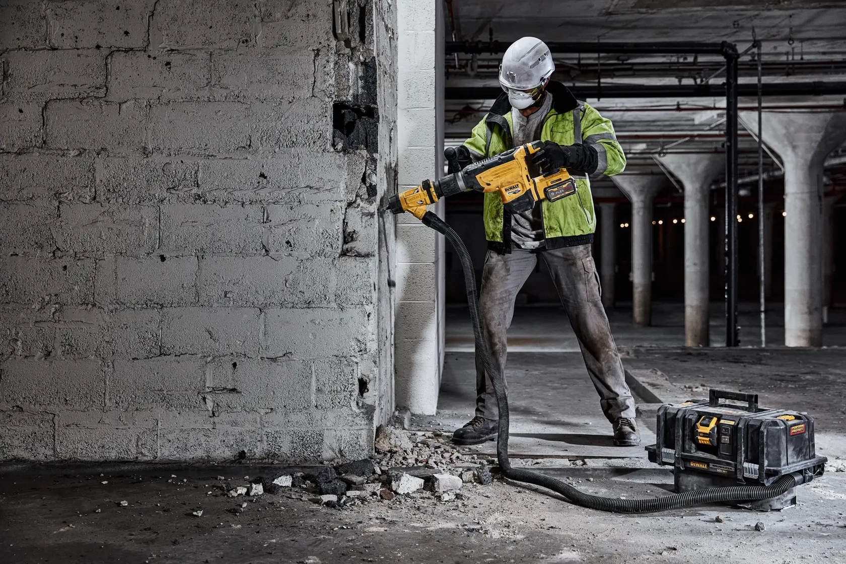 A construction worker wearing safety gear uses a yellow DEWALT cordless rotary hammer drill on a concrete wall. Debris is scattered on the ground, and a DEWALT tool case with attached hose is positioned nearby.