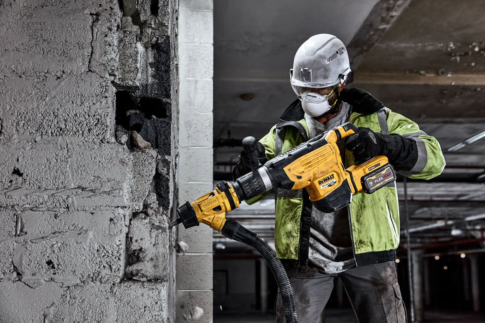 A construction worker wearing a safety helmet and gloves is using a yellow DEWALT cordless rotary hammer to drill into a concrete wall. The power tool is connected to a dust extraction system. The scene appears to be inside a building under construction or renovation.