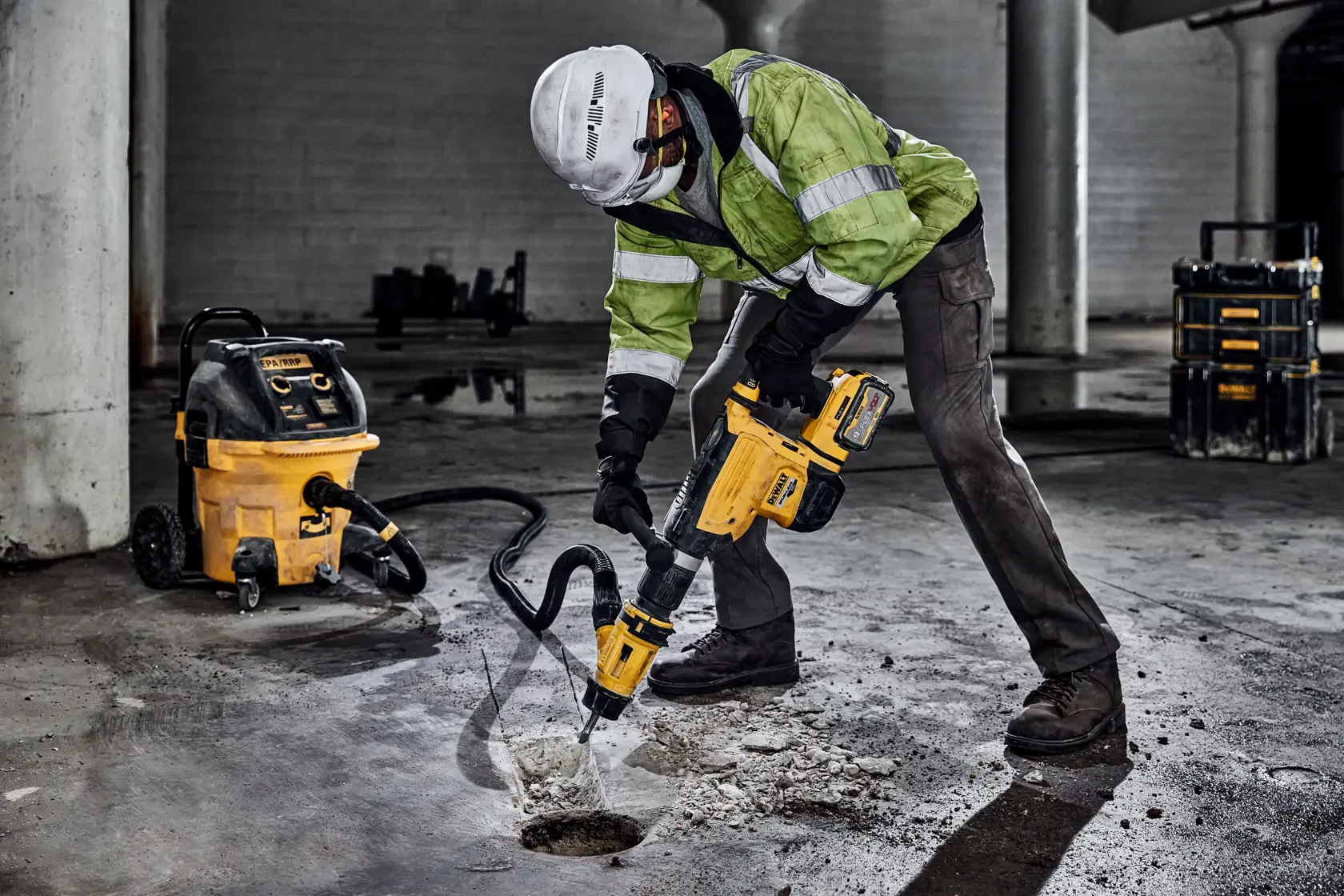 A worker wearing safety gear operates a DEWALT hammer drill to break up concrete on a construction site. A DEWALT vacuum is connected to the tool for dust extraction.
