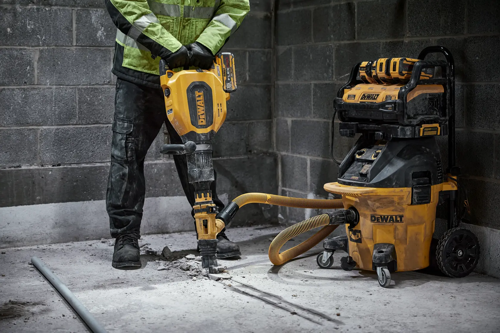 A person in workwear is operating a DEWALT demolition hammer, breaking up a concrete floor in an industrial setting. A DEWALT dust extractor is attached to the demolition hammer to manage debris.