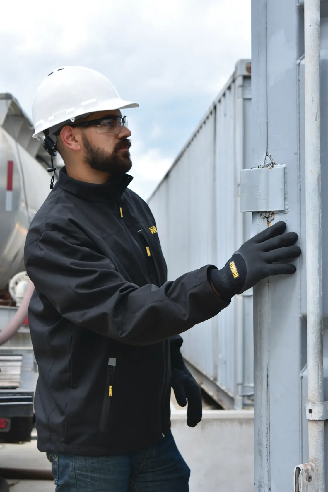A person wearing a black DEWALT jacket, black gloves, and a white hard hat is standing outdoors near an industrial structure, reaching toward a metal gate or door. The person's face is blurred for privacy.