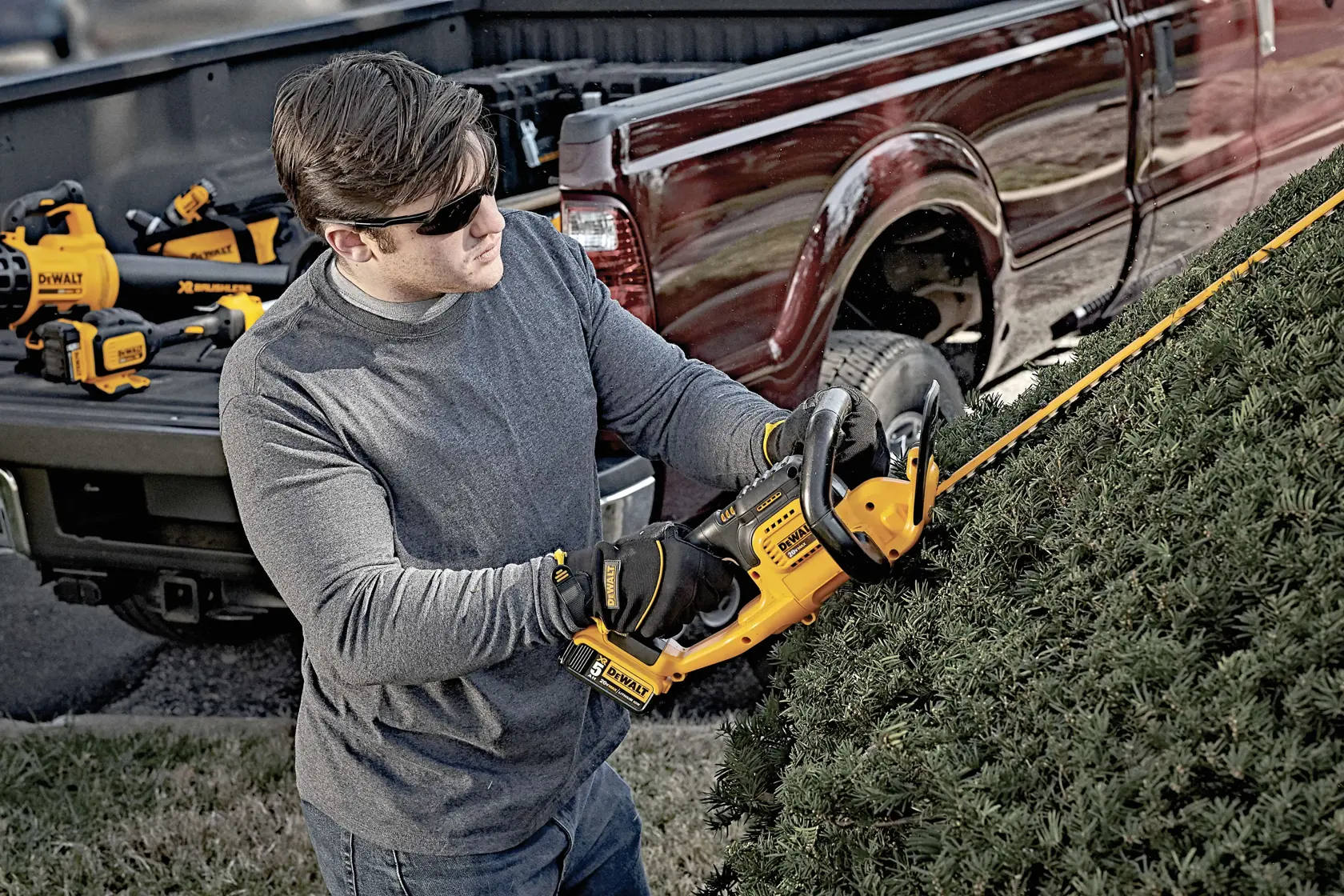 A person wearing gloves and a gray shirt uses a DEWALT cordless hedge trimmer to trim bushes next to a red pickup truck. Several DEWALT power tools are visible in the truck bed.