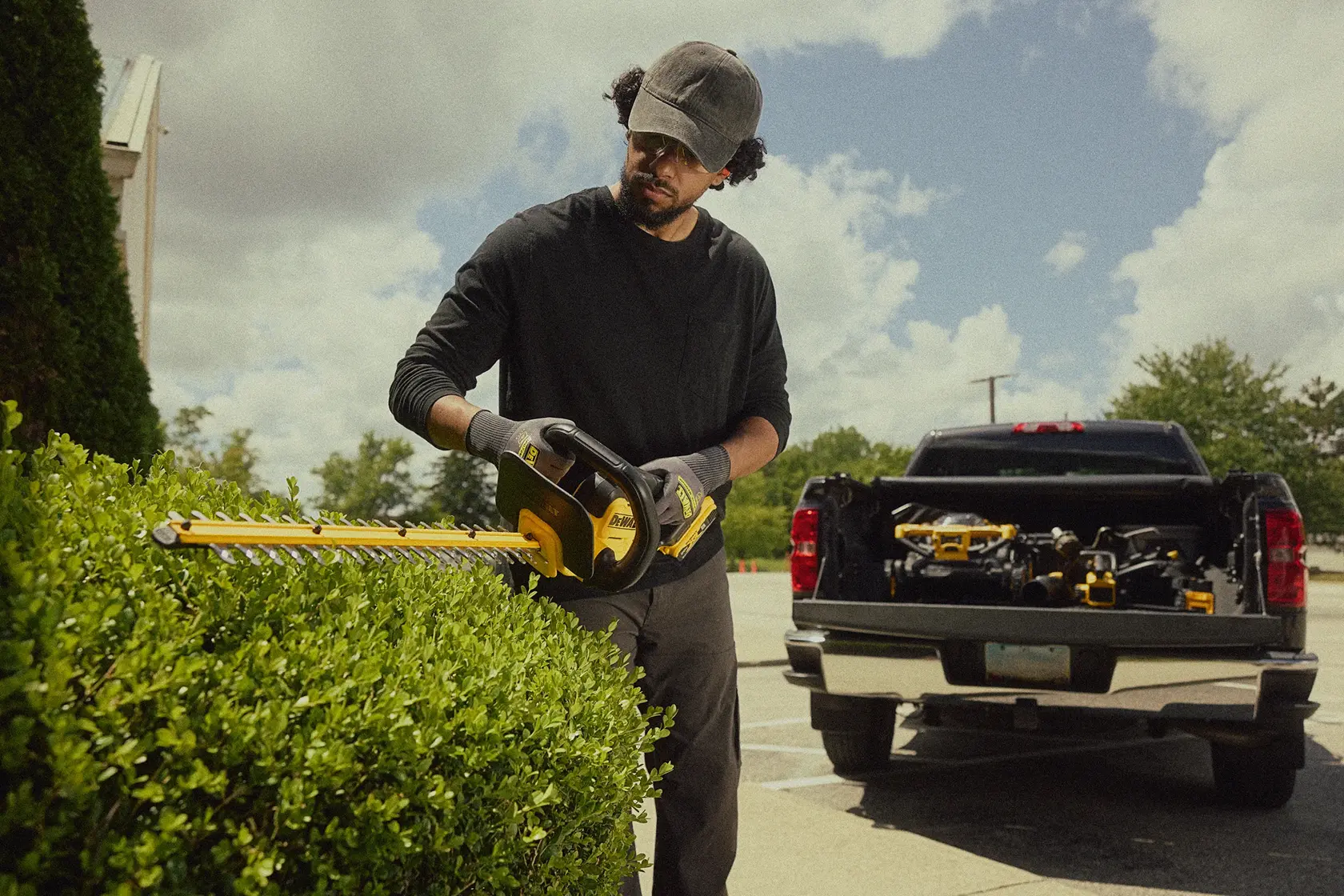 A person using a yellow DEWALT cordless hedge trimmer to trim bushes outdoors. The person is wearing gloves and a cap, and a pickup truck filled with DEWALT tools is parked nearby.