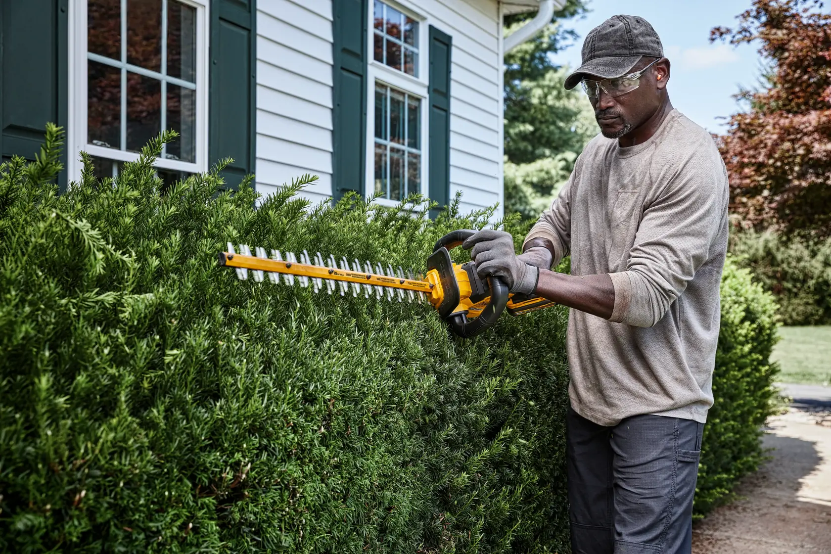 A person wearing gloves and a hat is using a DEWALT hedge trimmer to trim bushes outside a house with white siding and green shutters.