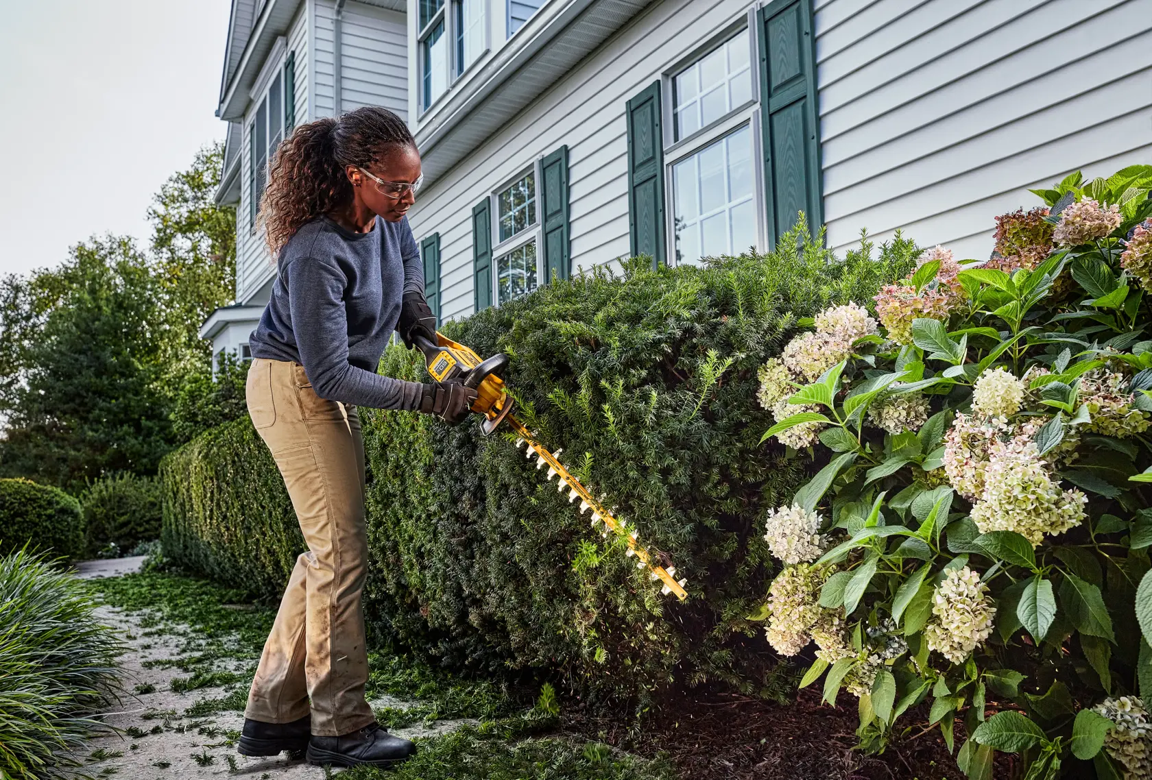 A person is using a DEWALT cordless hedge trimmer to trim bushes in front of a house with white siding and green shutters. The hedge trimmer is yellow and black, and the area is landscaped with shrubs and flowering plants.