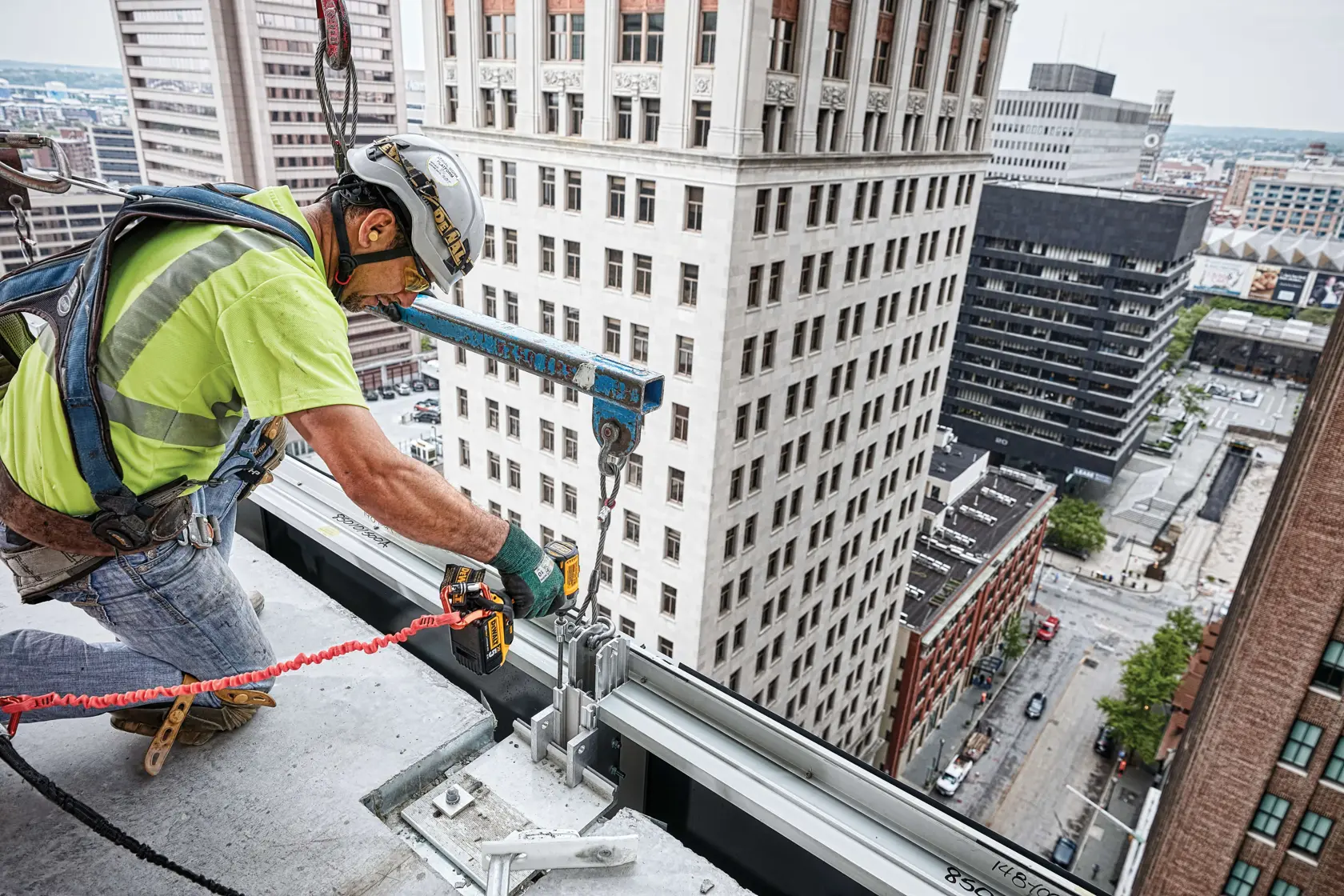A construction worker wearing safety gear is using a DEWALT power tool to work on metal framing at the edge of a high-rise building, with city skyscrapers visible in the background.