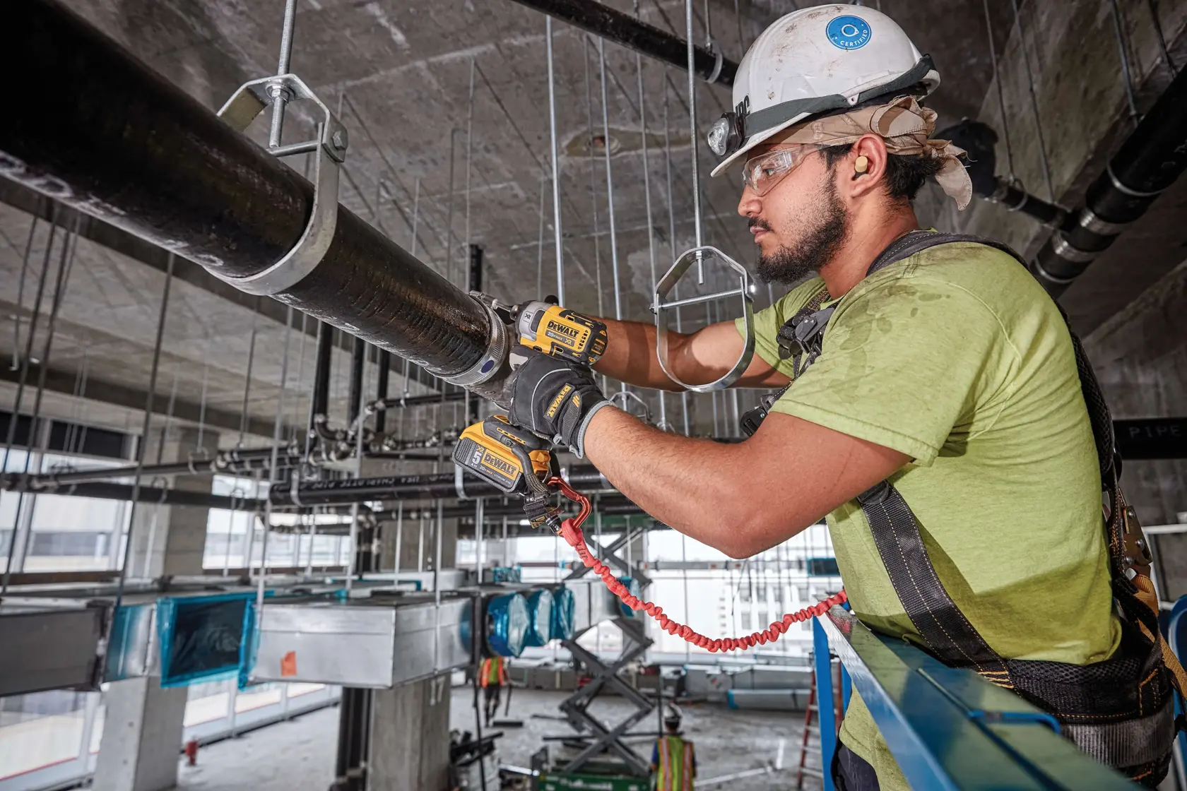 A construction worker wearing a safety harness and helmet is using DEWALT tools to secure or work on a large suspended pipe in an industrial setting.