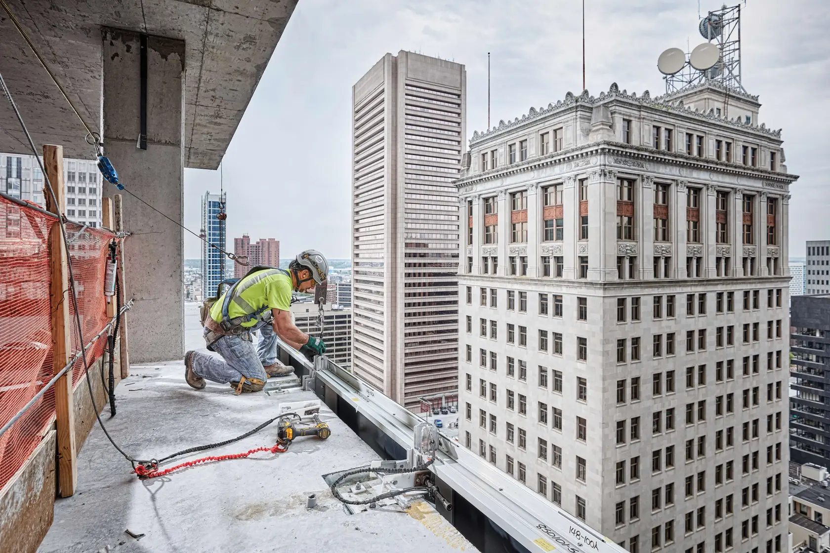 Construction worker in safety gear using DEWALT tools on the edge of a high-rise building under construction, with city buildings visible in the background.