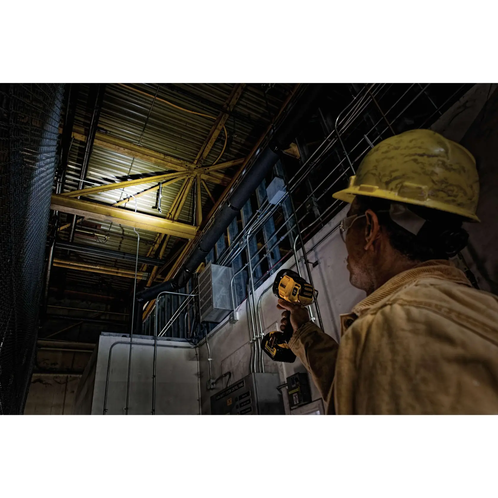 A worker wearing a yellow hard hat and jacket is holding a DEWALT DCL043 spotlight, shining it up toward the ceiling of an industrial building, illuminating pipes and metal beams.