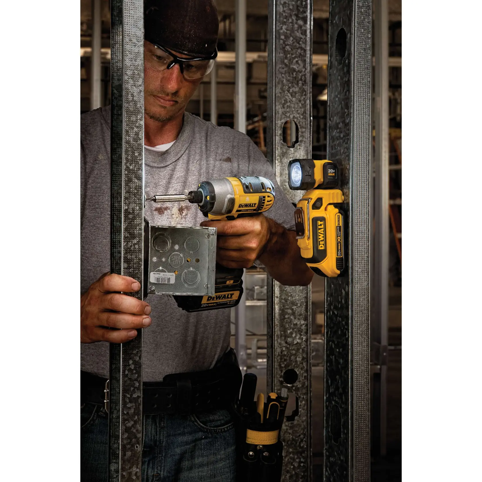 A person using a DEWALT cordless drill on a metal electrical box, with a DEWALT DCL044 LED work light attached to a metal frame. The scene appears to be at a construction site with the person's face blurred for privacy.