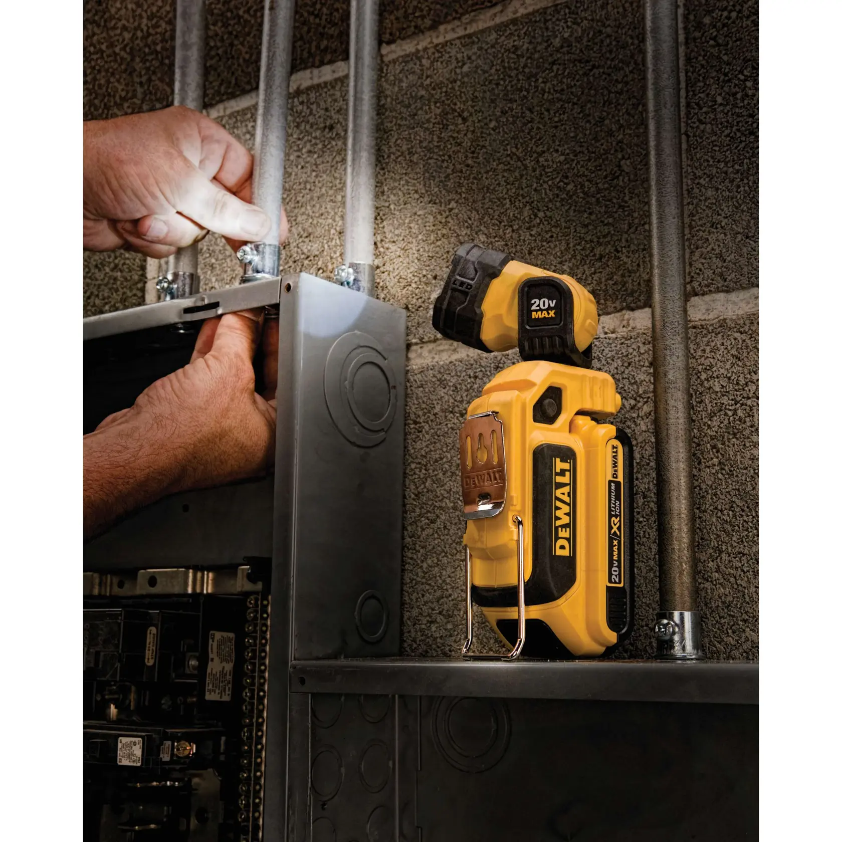 A DEWALT portable work light model DCL044 is shown illuminating a workspace inside an electrical panel. A person's hands are working on wiring, and the light is directed toward the panel to provide visibility.