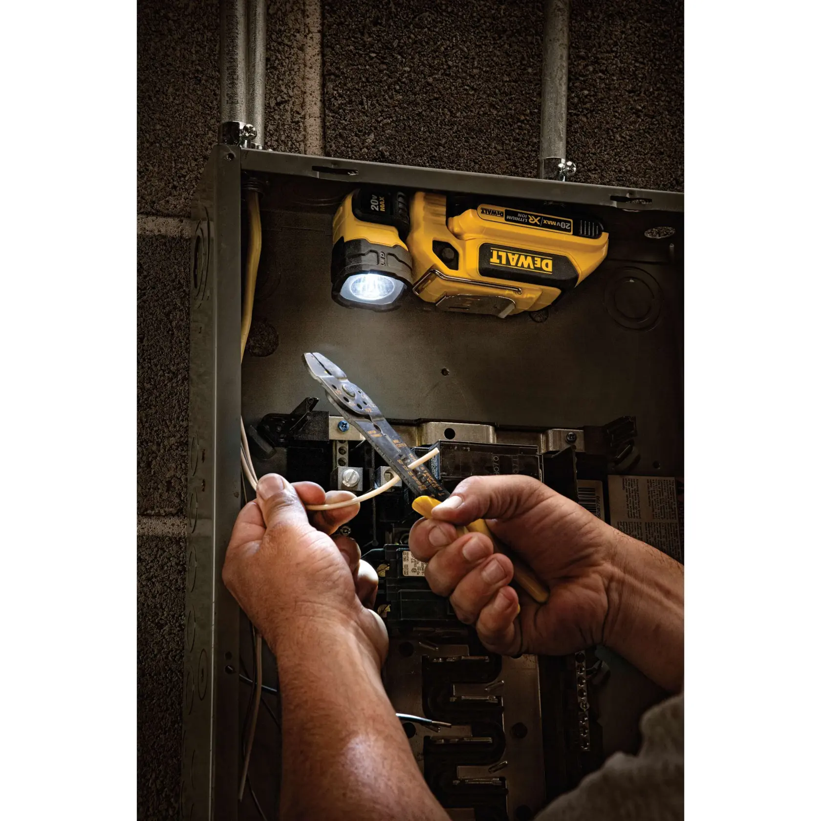 A DEWALT portable LED work light (model DCL044) illuminates the inside of an electrical panel while hands use pliers to strip wires.
