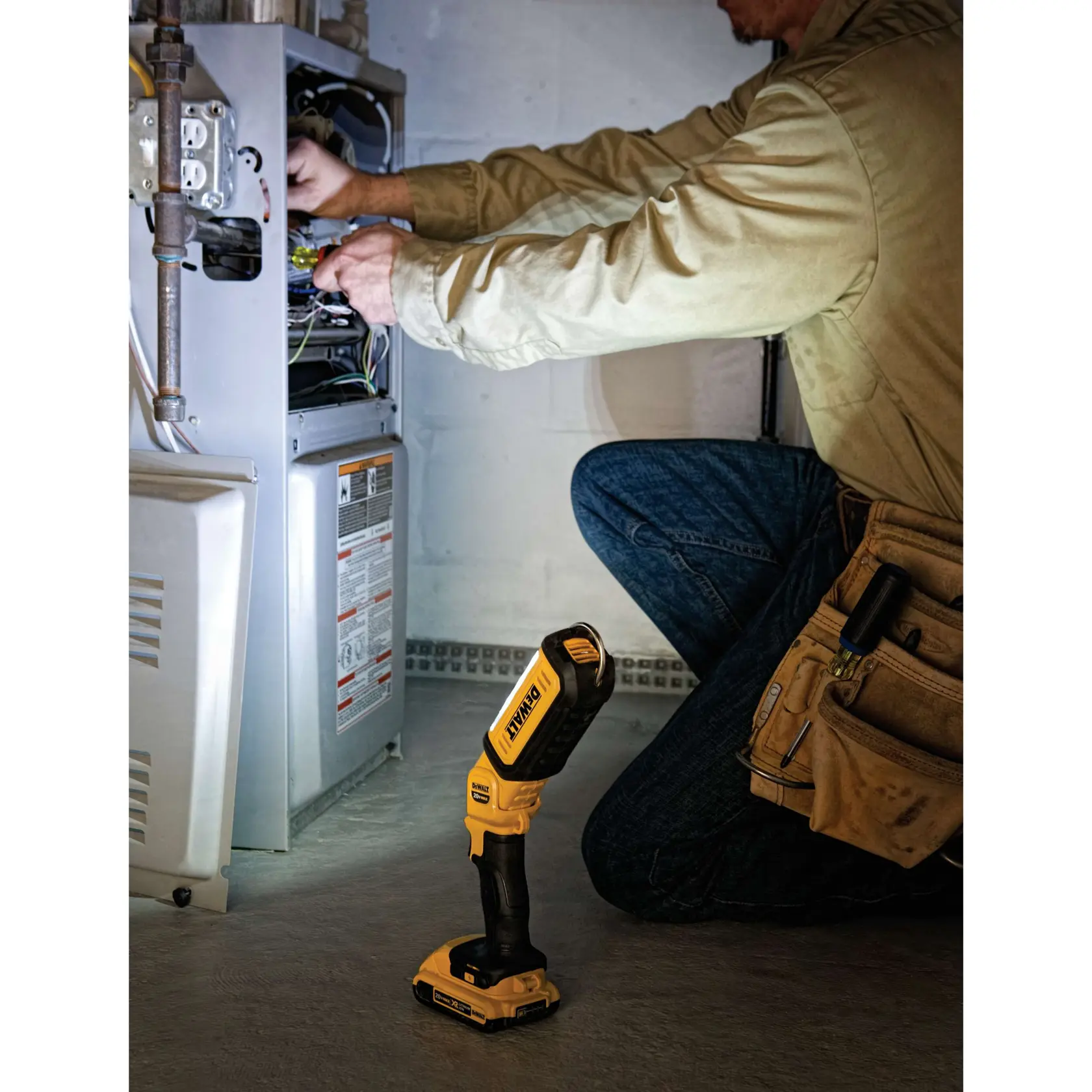 A person is kneeling and working on an open electrical unit, using tools. On the floor is a DEWALT DCL050 portable LED work light, illuminating the workspace.