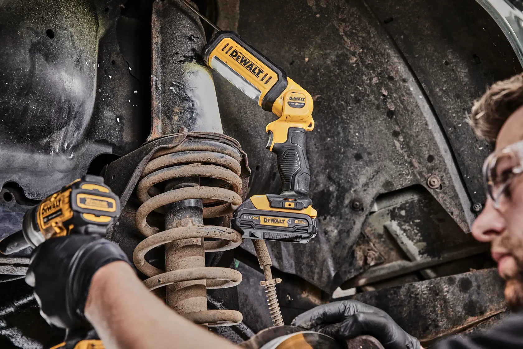 A DEWALT DCL050 LED work light is being used to illuminate an automotive suspension system while a person wearing safety glasses and gloves works with another DEWALT tool.