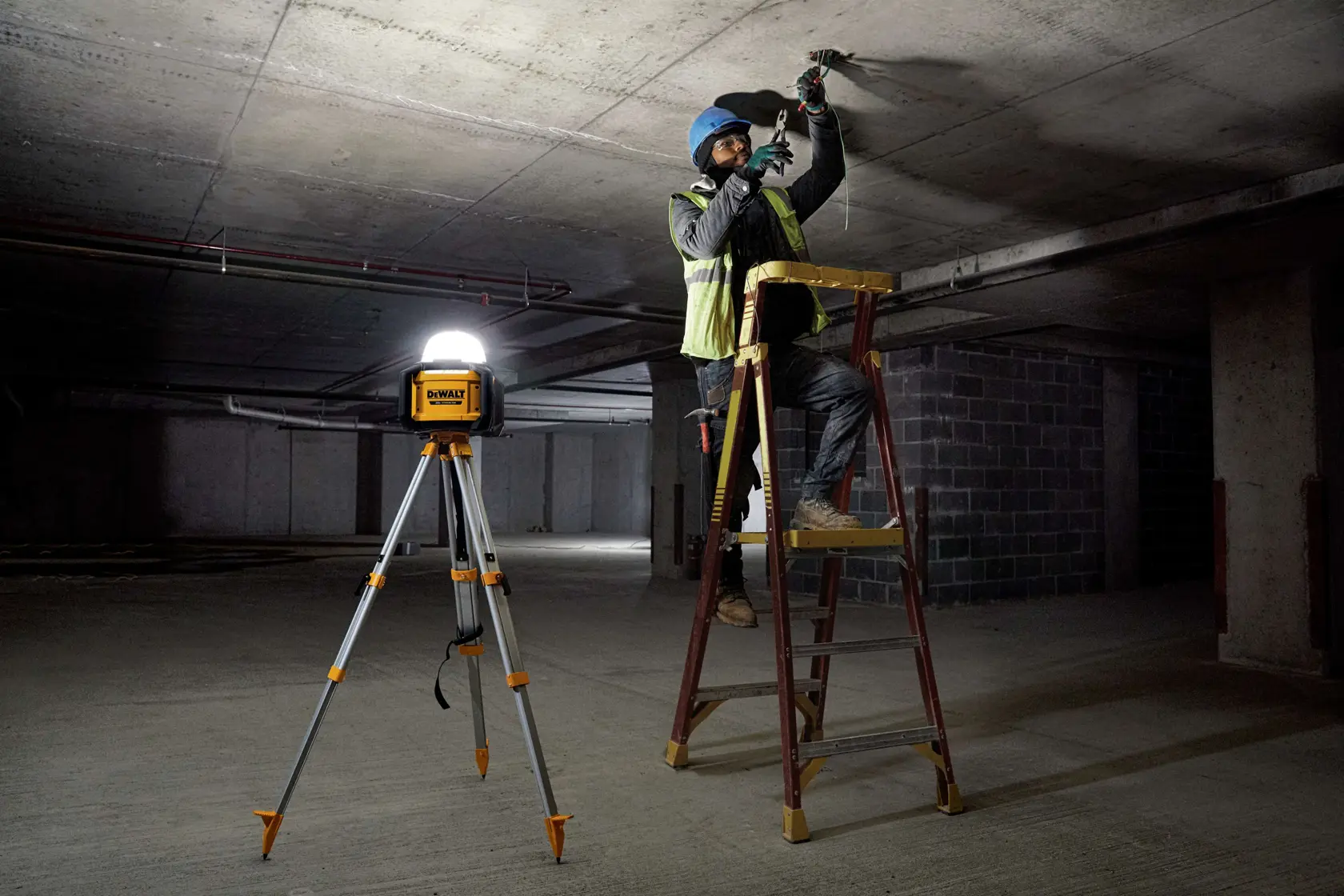 A DEWALT portable work light mounted on a tripod illuminates a dim construction site. Nearby, a worker wearing safety gear stands on a ladder and works on the ceiling.
