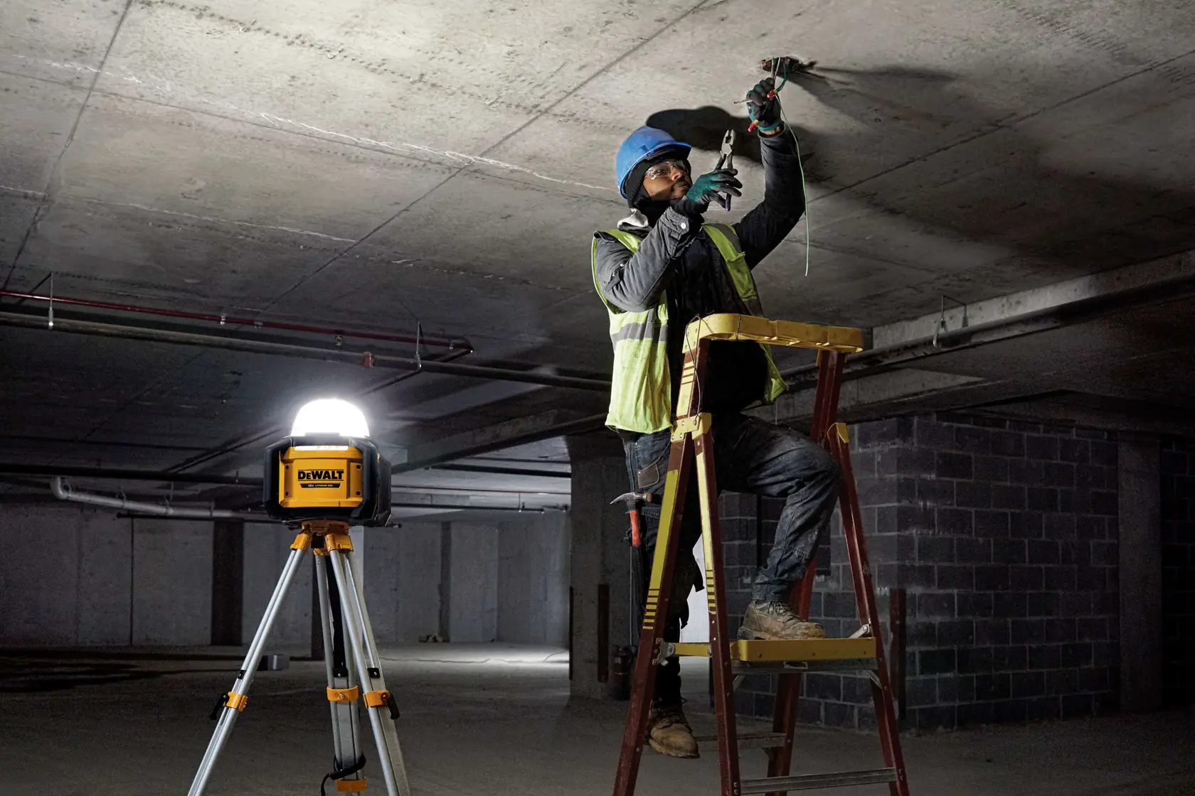 A DEWALT DCL074 work light on a tripod provides illumination for a construction worker on a ladder, working on the ceiling of an unfinished building.