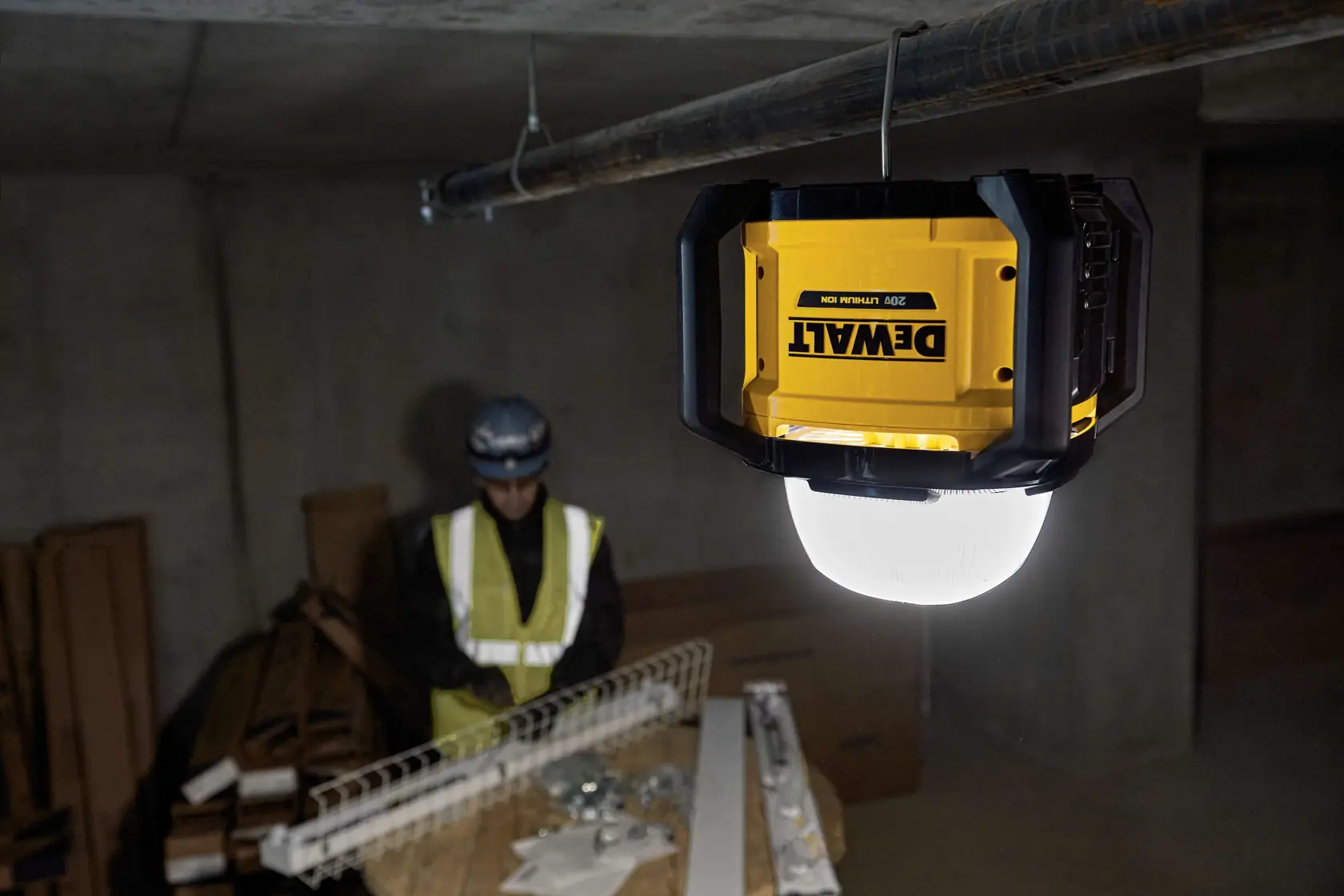 A DEWALT portable work light mounted on a tripod illuminates a dim construction site. Nearby, a worker wearing safety gear stands on a ladder and works on the ceiling.