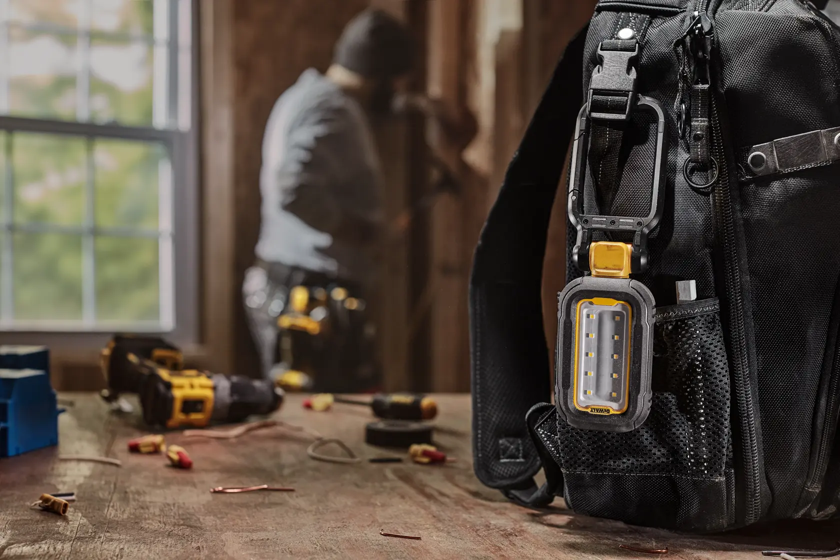 A DEWALT DCL182 portable LED work light attached to the strap of a black tool bag. The bag is on a wooden table with various tools and electrical connectors, while a person works in the blurred background.