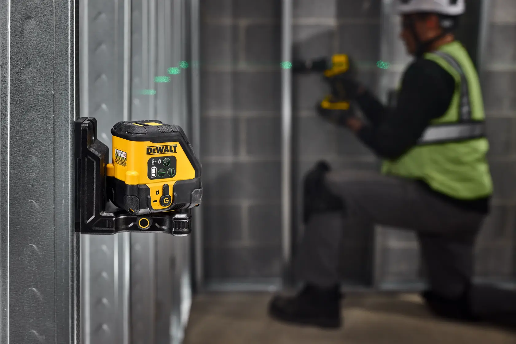 A DEWALT laser level tool mounted on a metal wall frame, projecting a green laser line across the room. In the background, a person wearing a safety helmet and reflective vest is working with another DEWALT tool, kneeling near the metal frame.
