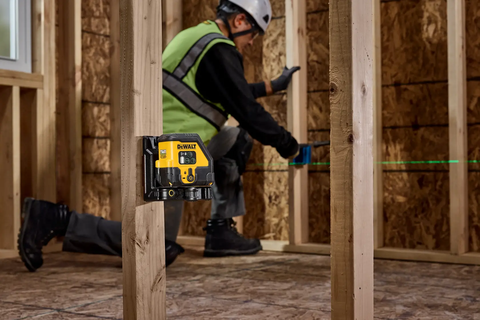 A DEWALT laser level mounted on a wooden stud emits a green laser line in a construction framing environment. A person wearing safety gear works in the background measuring or marking the wall.