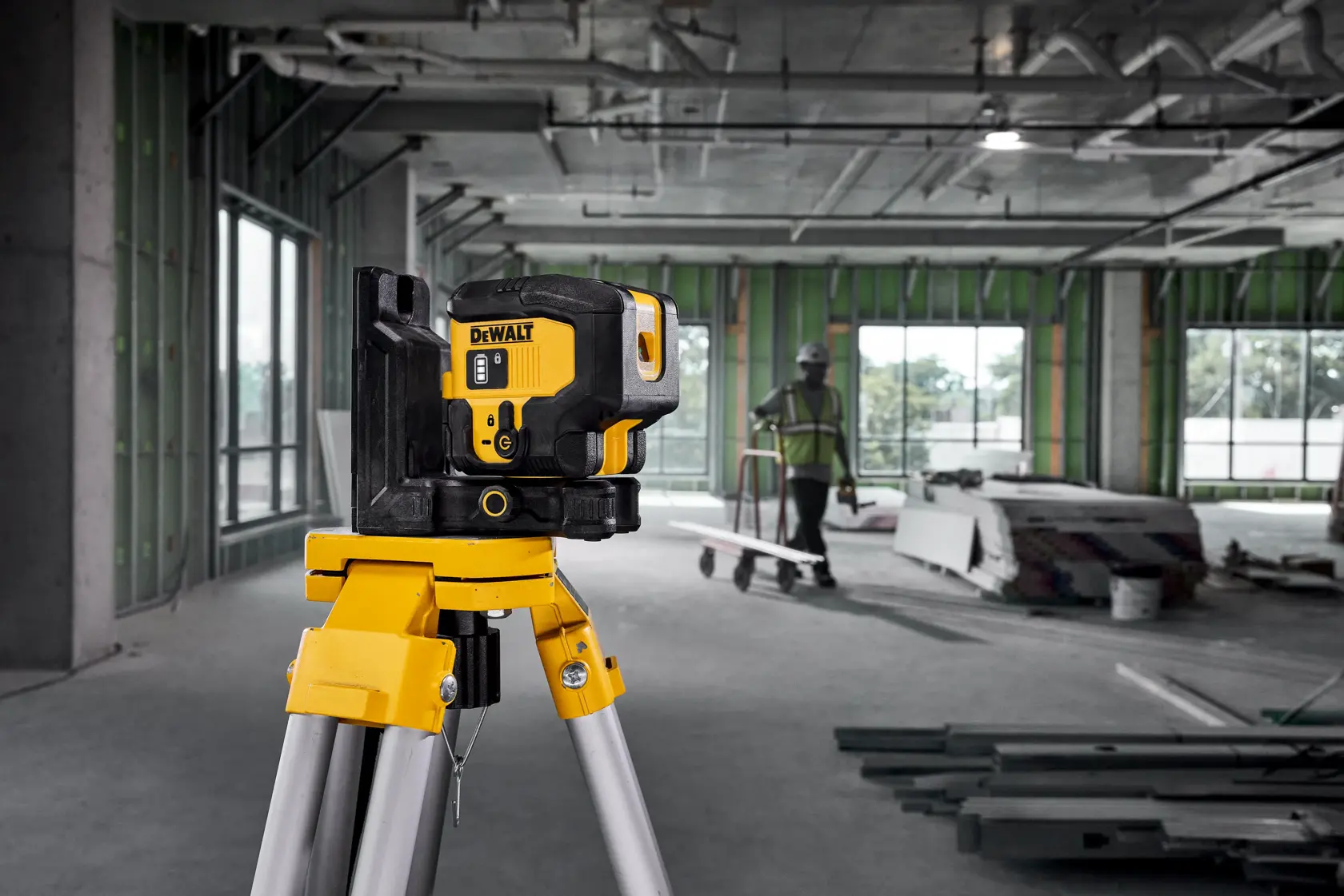 A DEWALT laser level mounted on a tripod is set up inside a construction site with unfinished walls and windows. Building materials and a person wearing a safety vest and helmet are visible in the background.