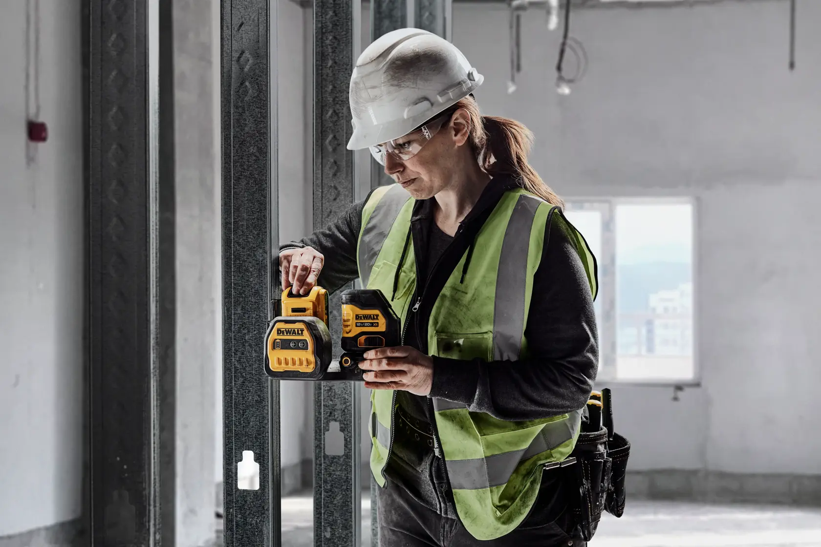 A person wearing a safety helmet and high-visibility vest is using a DEWALT tool in an unfinished construction setting. The DEWALT device is yellow and black, and the person appears to be adjusting or operating it.
