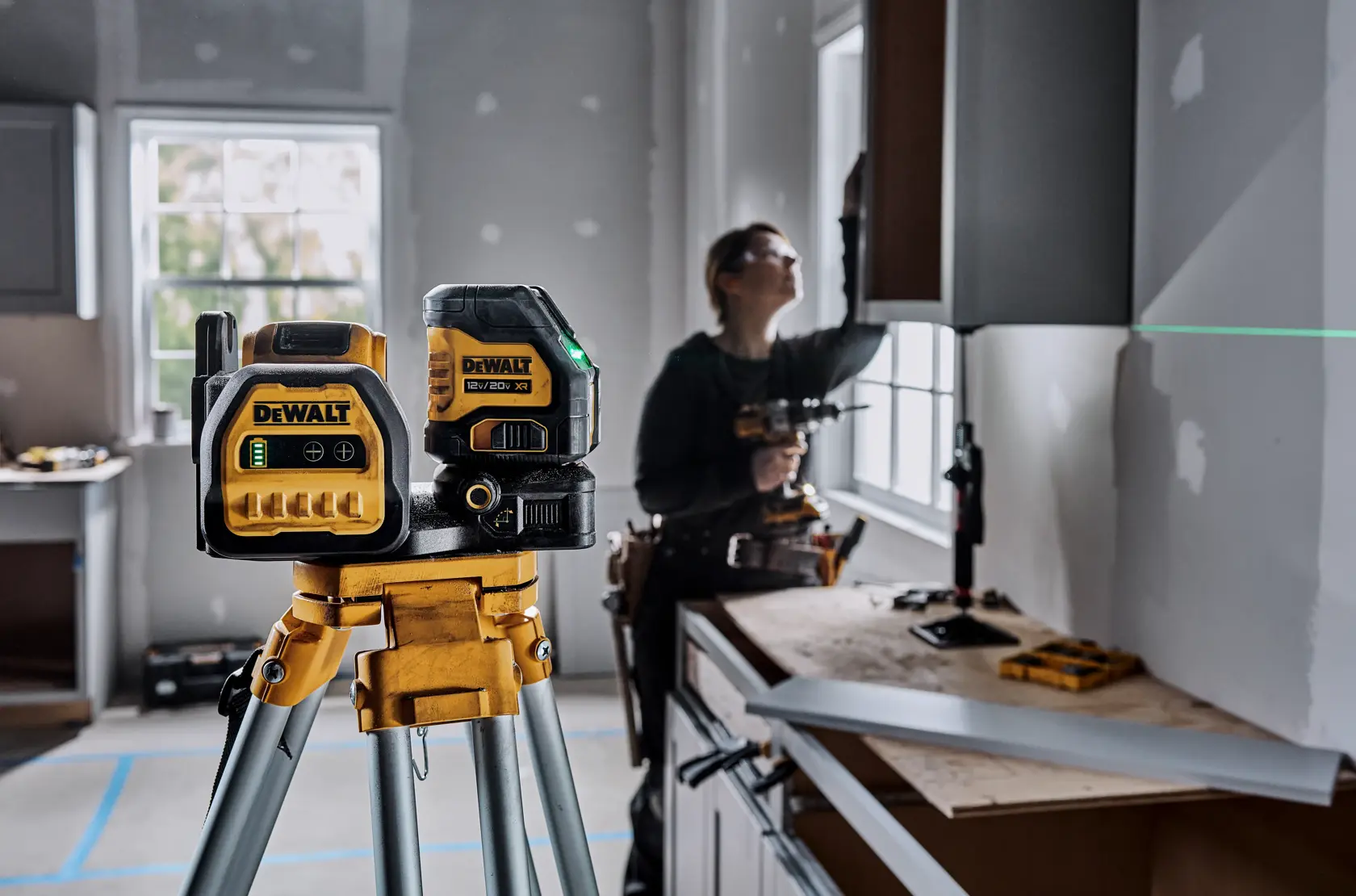 A DEWALT green laser level tool is mounted on a tripod in the foreground of a construction or renovation site, with a person working in the background installing cabinetry. The laser projects a green horizontal line across the wall, assisting in alignment.