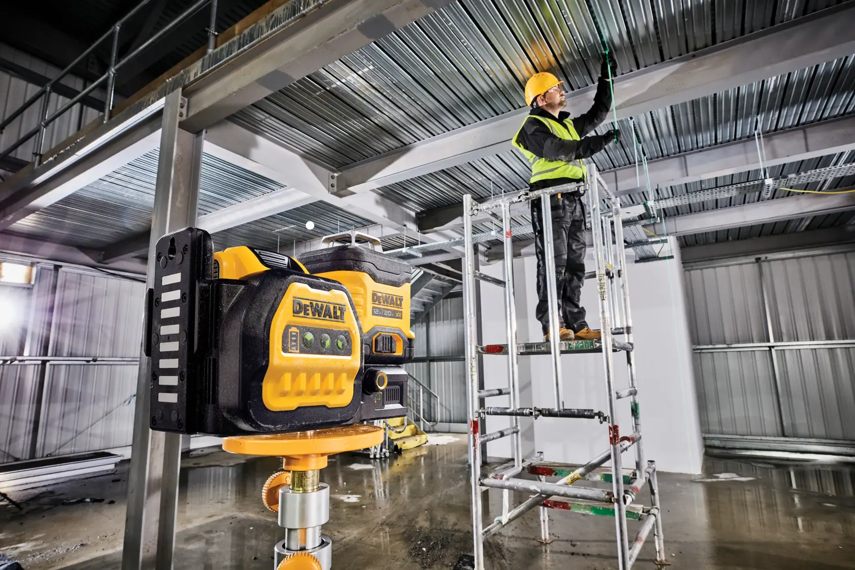 A DEWALT laser level device is mounted on a stand in a construction site, with a worker wearing safety gear operating on scaffolding in the background.