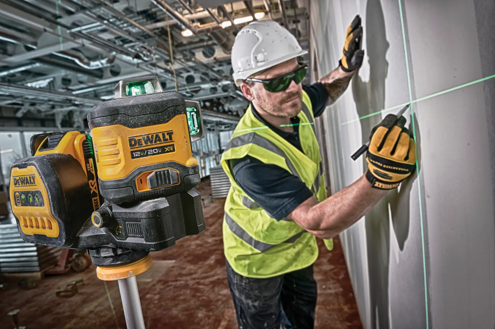 A DEWALT 12V/20V XR laser level device is set up on a tripod in the foreground, projecting green laser lines onto a wall. In the background, a construction worker wearing safety gear, including a helmet, gloves, and a high-visibility vest, is marking the wall using the laser lines for guidance.