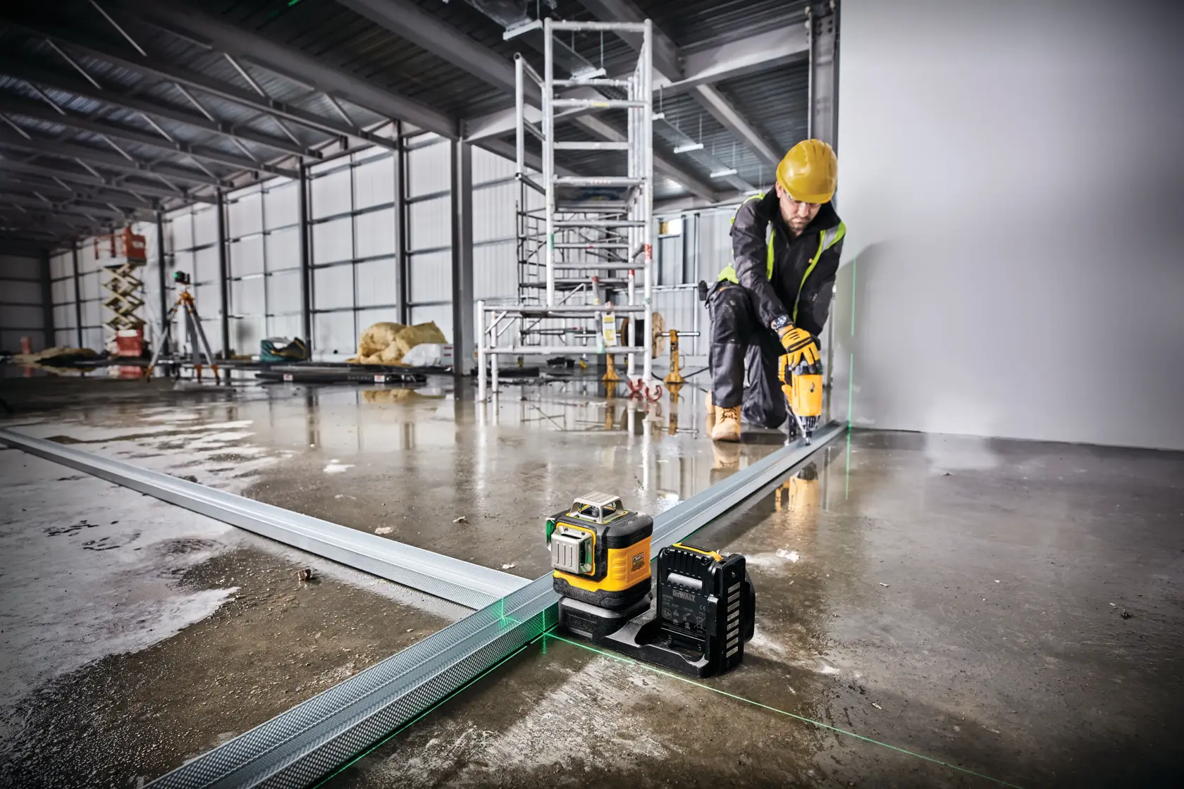 A DEWALT laser level tool is placed on the floor in a construction site, projecting green laser lines along metal tracks. A person wearing a yellow hard hat and safety gear is working in the background near the wall. The site has scaffolding and construction materials visible.