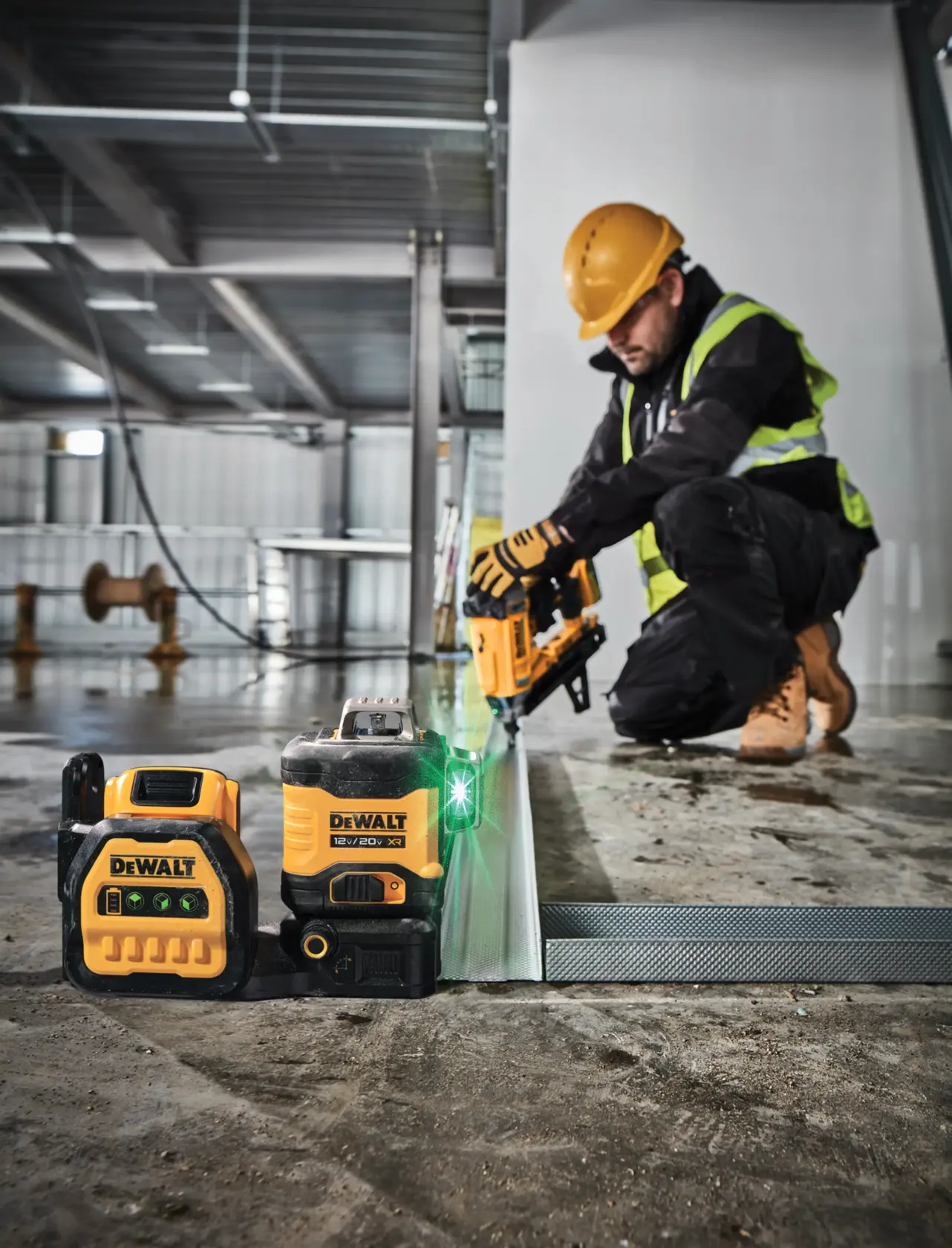 DEWALT laser level device and battery placed on a construction site floor with a construction worker in safety gear using a power tool in the background.